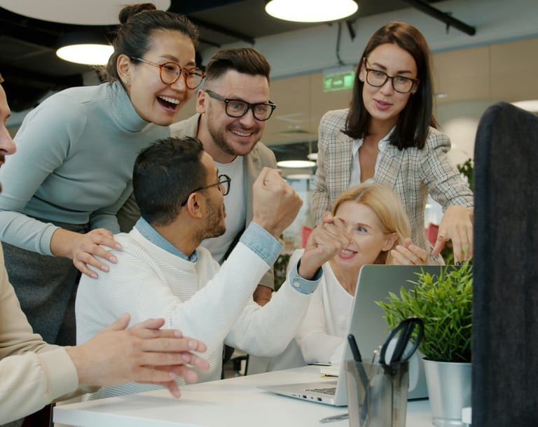 Diverse creative office team cheering and celebrating success while looking at a laptop screen together.