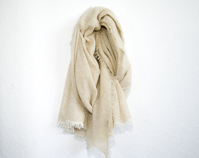 Cream pashmina shawl with fringed edges hanging on white wall, soft texture and cozy appearance.