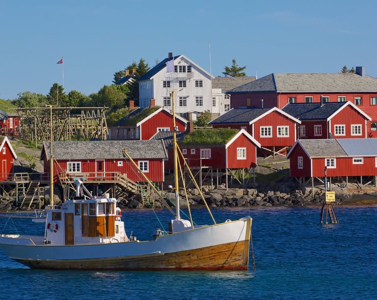 Fiskebåt foran røde rorbuer i Lofoten, sjø og bryggeanlegg i sollys.