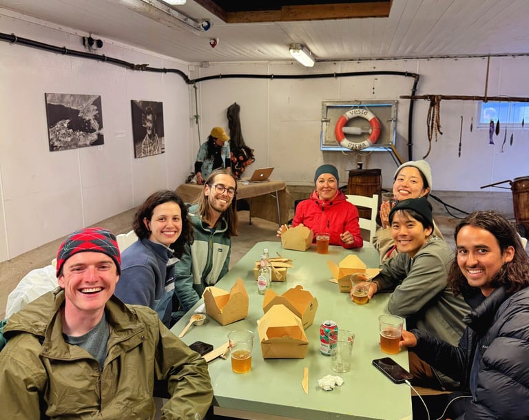 Gruppe gjester som spiser takeaway sammen ved langbord innendørs, matbokser og drikke på bordet.