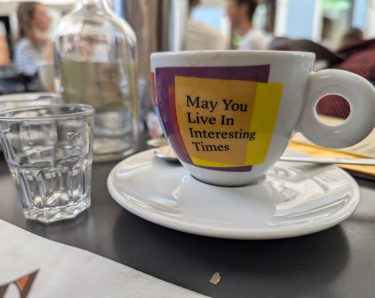 A coffee cup on a table, with the text 'May you live in interesting times' written on the cup.