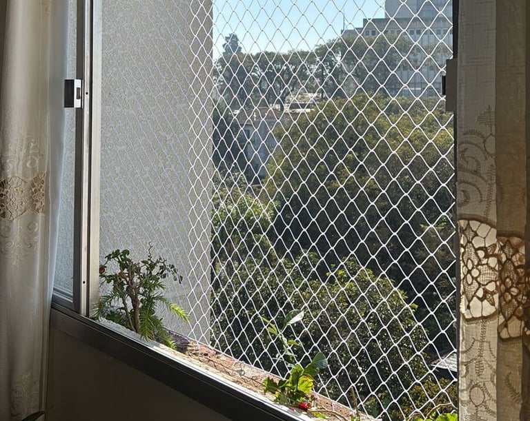 janela de apartamento vista por dentro com rede de proteção; cortinas claras nas laterais, plantas no parapeito e, ao fundo,