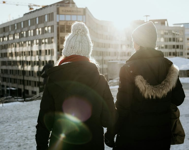Two women in winter coats and beanies walking through a snowy city at sunset.