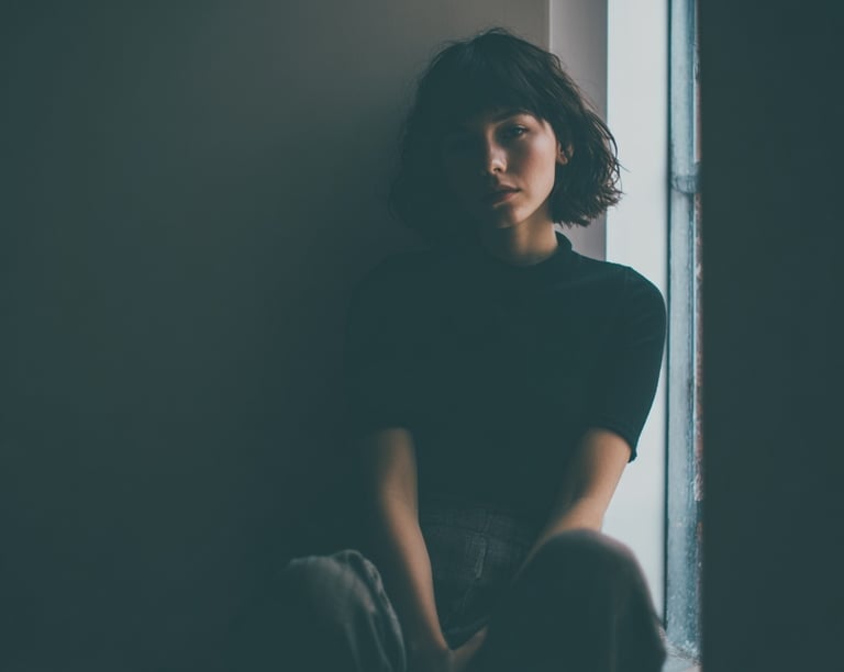 Moody portrait of a woman with short hair sitting by a window in a dimly lit room.