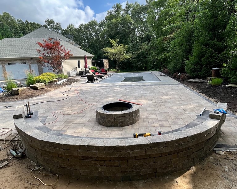 Custom paver patio installation with stone retaining wall, fire pit, and pool deck in a backyard.