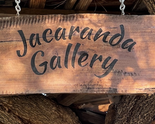 Rustic carved wooden sign for Jacaranda Gallery hanging by metal chains from a tree.
