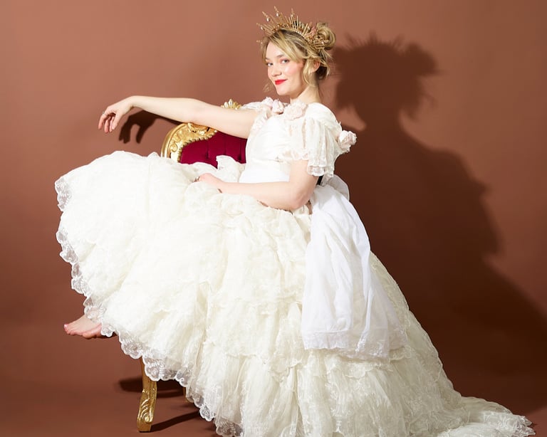 A woman in a ruffled white ballgown and gold crown posing on a vintage velvet chair.