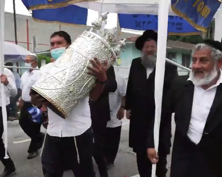Jewish men carry a silver Torah scroll during a festive outdoor parade under a blue and white canopy.