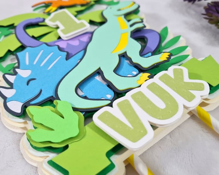 Colorful paper cake topper featuring a cartoon dinosaurs theme with the number "1" at the center. Th