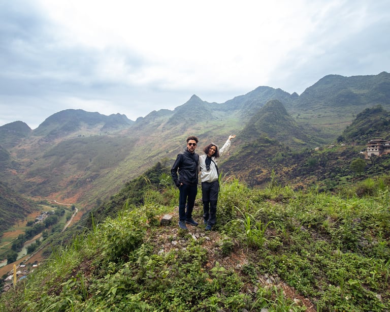 A couple standing on a grassy mountain edge posing for the camera. in the background are the beautiful Ha Giang mountains. 