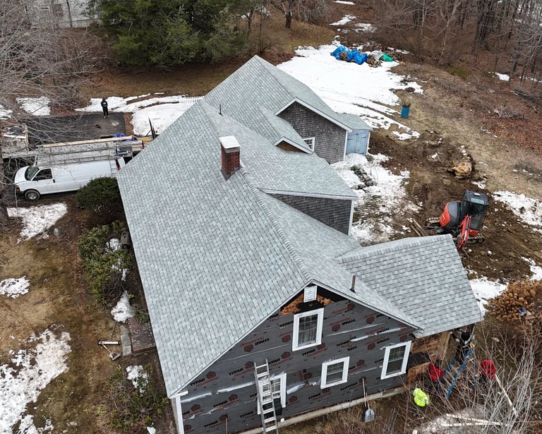 Aerial view of a residential roof replacement and home renovation project with grey shingles and excavating equipment.
