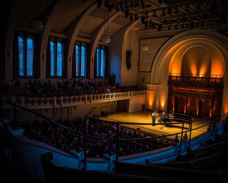 Violinist and pianist perform a classical music concert for an audience in a historic grand hall.