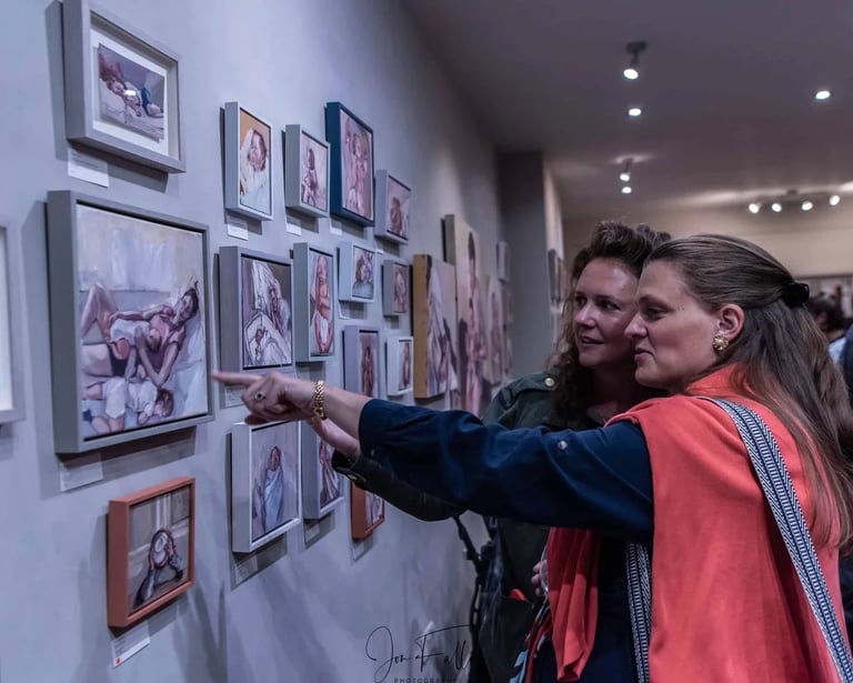Two women looking at a gallery wall of framed contemporary portrait paintings in an art exhibition.