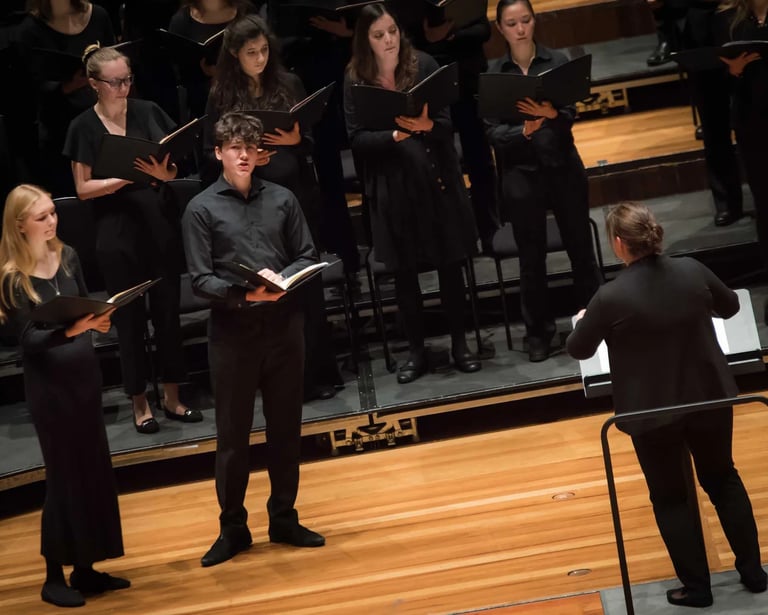 Professional choir singers in black formal attire performing a choral concert with a conductor on stage.