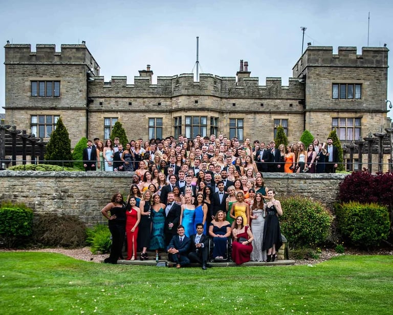 A large group of graduates in formal attire posing for a photo in front of a historic castle venue.