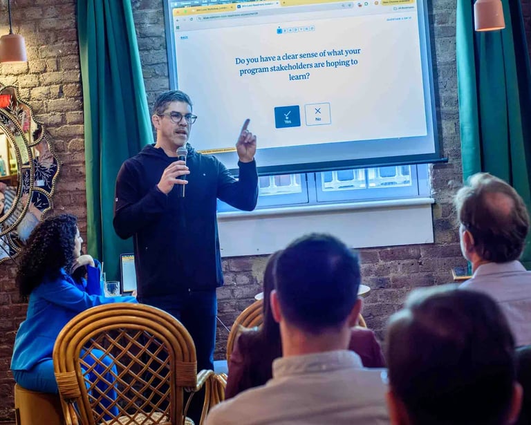 A man presents a question about program stakeholders to an audience, using a projector in a dimly lit room during a business