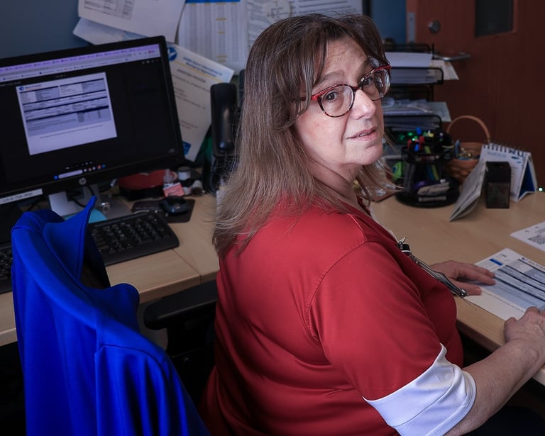 A female office professional wearing glasses and a red shirt reviews documents at her computer desk.