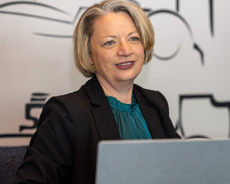A smiling woman with short blonde hair and glasses, wearing a dark blazer over a teal top, sits at a desk with a laptop.