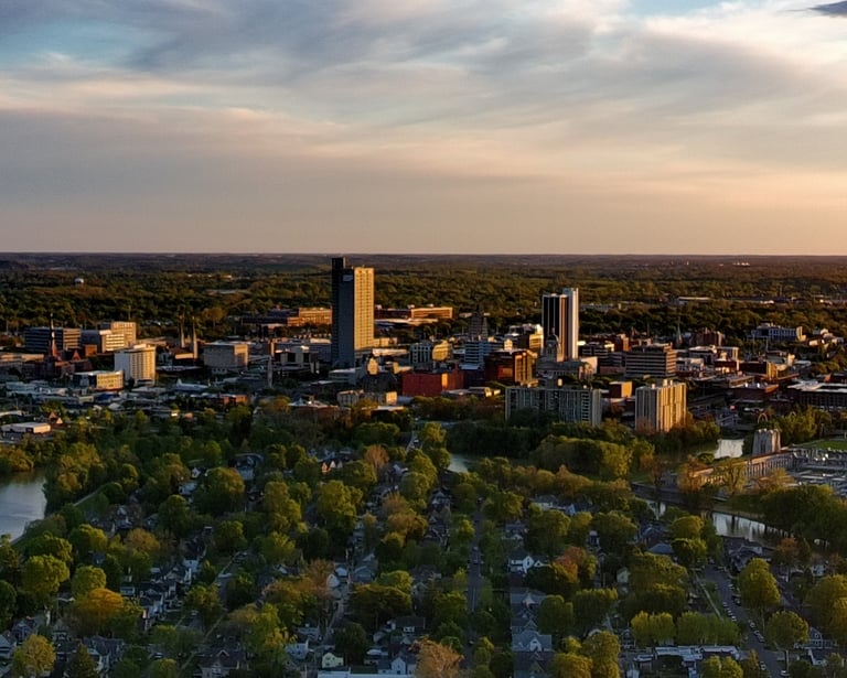 Aerial view of Fort Wayne Indiana skyline at sunset with St. Joseph River and lush green trees.