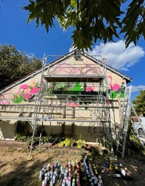 une grange en taule avec un échafaudage et une fresque avec des flamands roses et des plantes