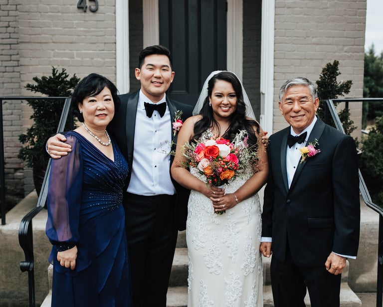 Smiling bride and groom posing with parents in formal wedding attire outside a brick house.