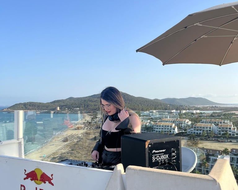 A female DJ performs at a rooftop beach club overlooking the coast with a Pioneer speaker and Red Bull booth.