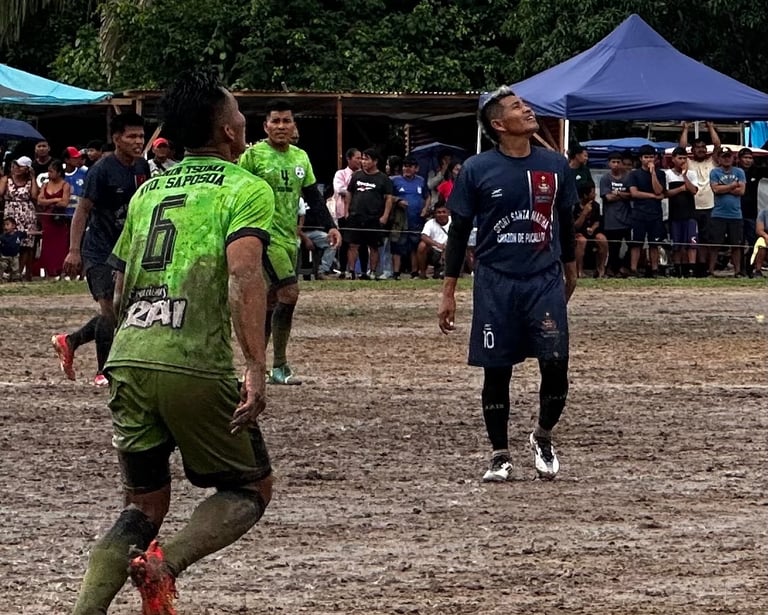 Soccer players competing on a muddy field during a local football match in a rural area.
