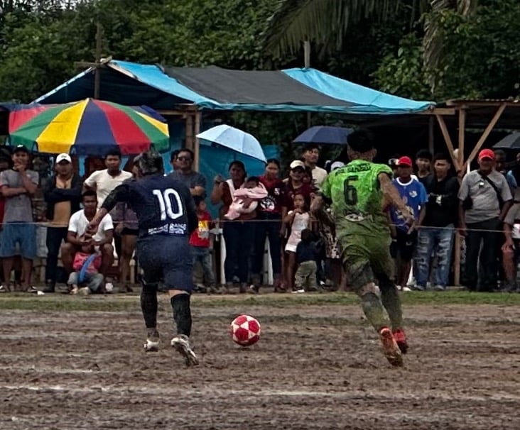Soccer players competing on a muddy field during a local football match with spectators in the background.