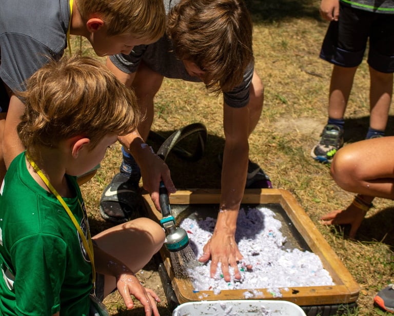 Teacher using a hose to spray shredded paper onto a screen with kids surrounding.
