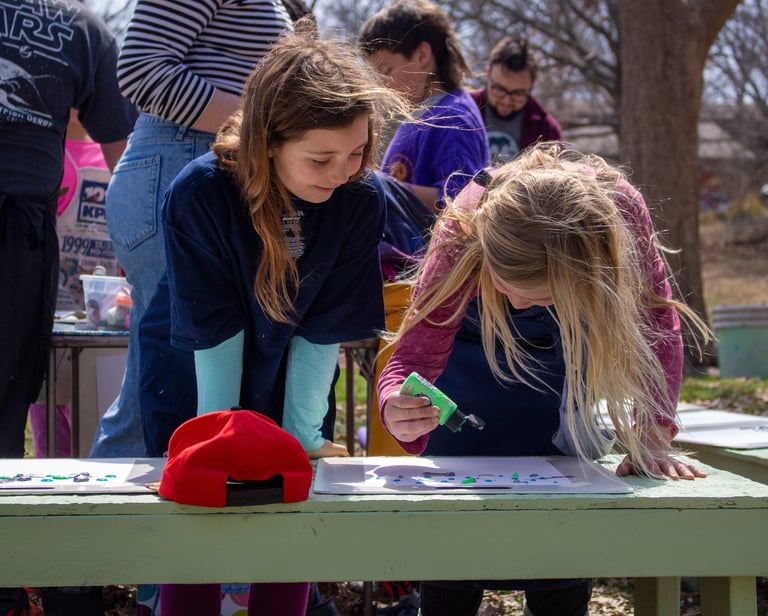 Two young girls smiling while standing over papers dripping paint onto them.