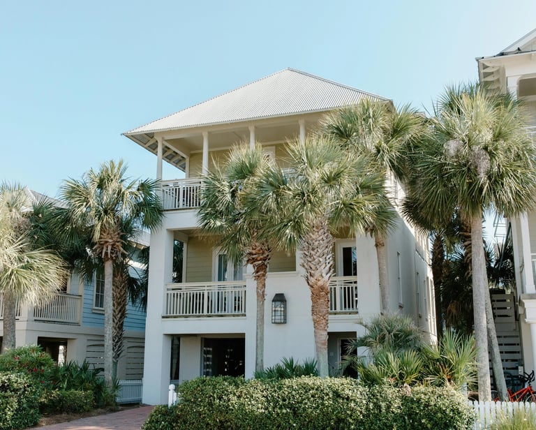 Luxury three-story beach house rental with palm trees and balconies under a clear blue sky.