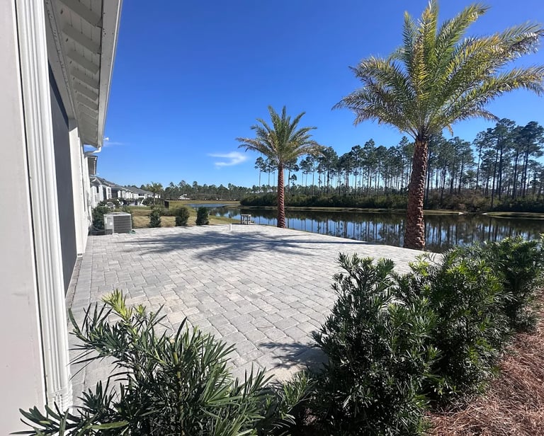 A large stone paver patio overlooking a scenic lake with palm trees and a clear blue sky.