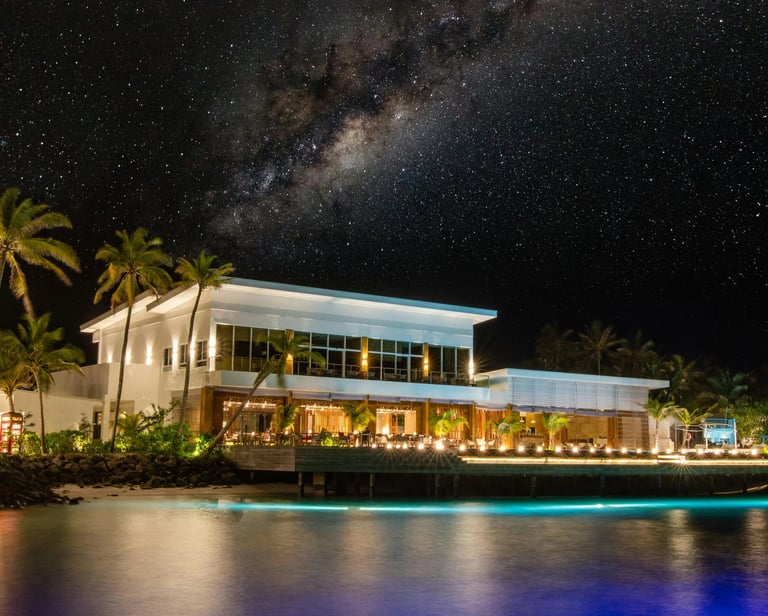Luxury tropical resort at night featuring the Milky Way galaxy above a lit beachfront restaurant and palm trees.