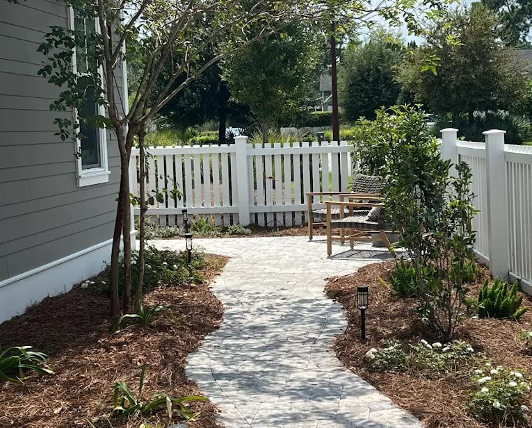Curving stone paver walkway in a backyard garden with pine straw mulch and a white picket fence.