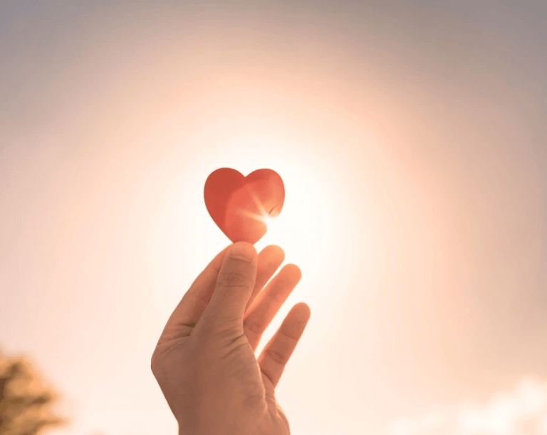 Une personne montre un cœur rouge devant le soleil, symbolisant l'amour authentique rayonnant.