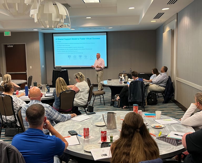 A presenter leads a business seminar on virtual success and shared support models for a group of professionals seated at