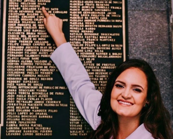 Dra Bruna Melchior de jaleco branco apontando seu nome em placa de metal do Hospital das Clinicas da USP de Ribeirao Preto