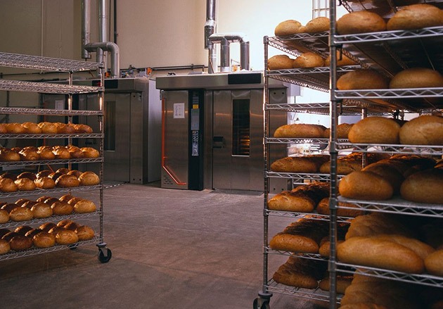 Freshly baked bread loaves cooling on metal racks in a commercial industrial bakery kitchen.