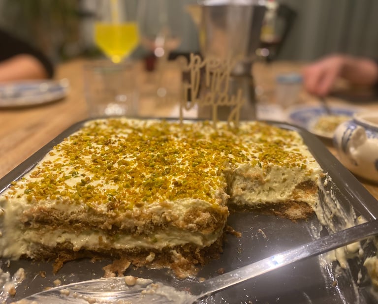 Homemade pistachio tiramisu cake topped with crushed nuts and a happy birthday sign on a dining table.