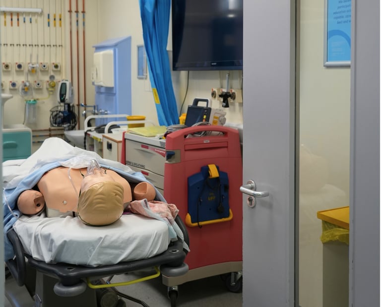 Medical simulation mannequin on a stretcher in a critical care training room with hospital equipment.
