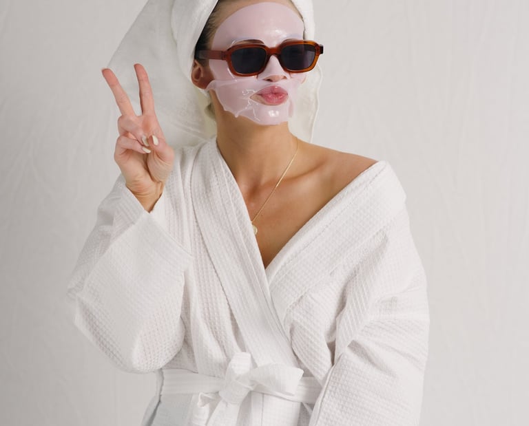 Woman in a white bathrobe and towel wrap wearing a pink sheet face mask and sunglasses while making a peace sign.