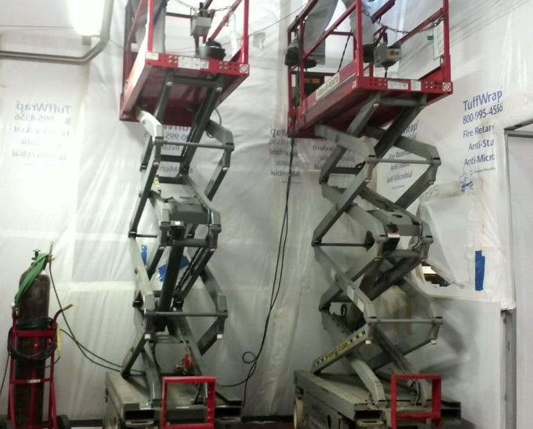 Two construction workers on scissor lifts performing pipe installation in dust containment area.