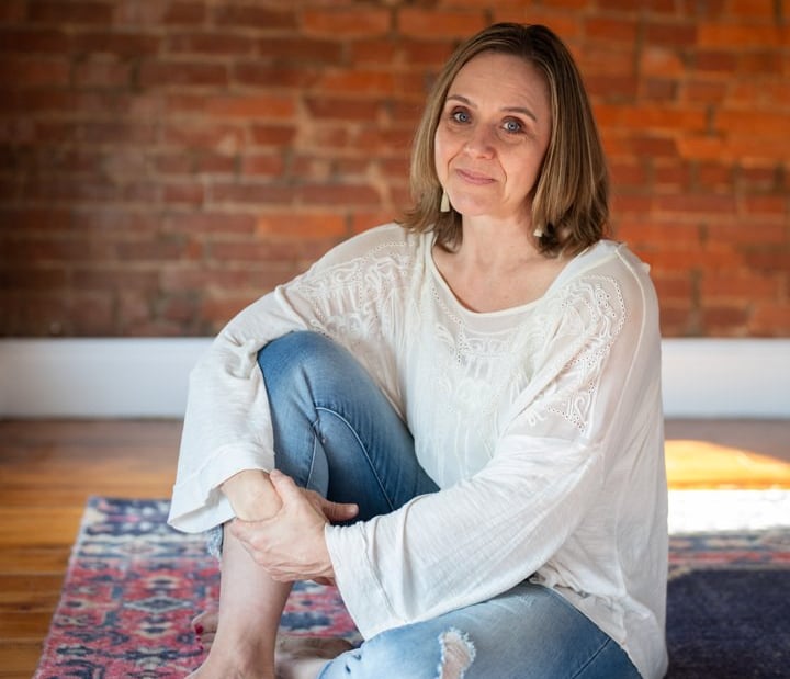 Woman sitting indoors on a rug in front of a brick wall.