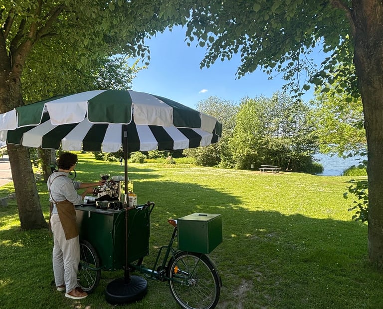 Mobiles dunkelgrünes Kaffeefahrrad mit grün-weißem Schirm am Seeufer.