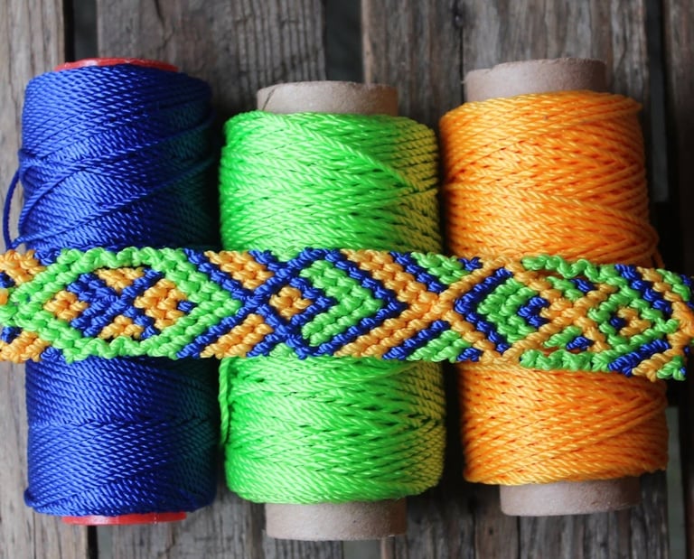 Pulsera  tejida a mano con rombos en azul, verde y naranja, presentada sobre carretes de hilo.