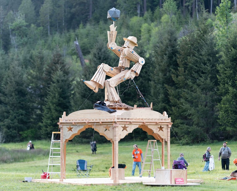 A large wooden figure sculpture atop a gazebo in a forest clearing for an outdoor art installation.