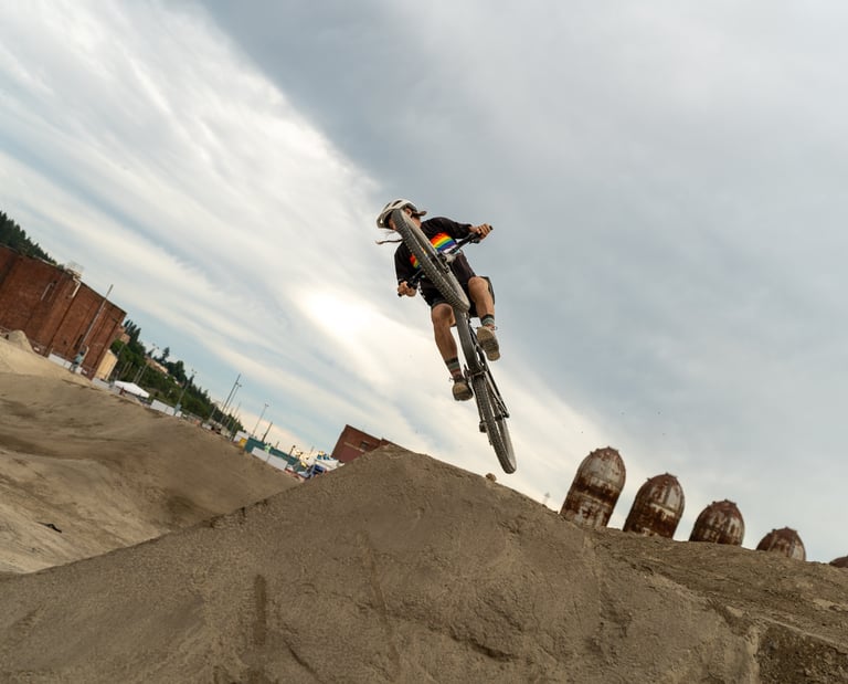 A mountain biker catches air off a dirt jump at an industrial bike park under a cloudy sky.