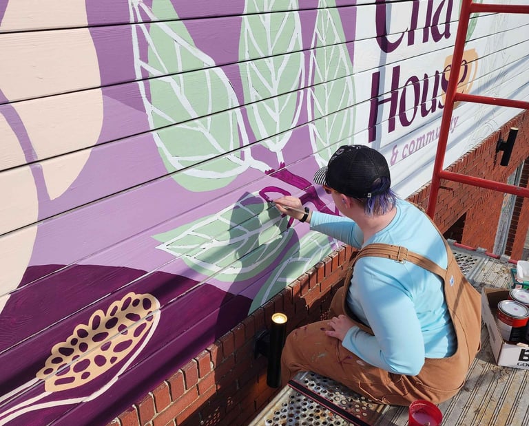 Artist painting a colorful tea leaf mural on an exterior building wall using purple and green paint.