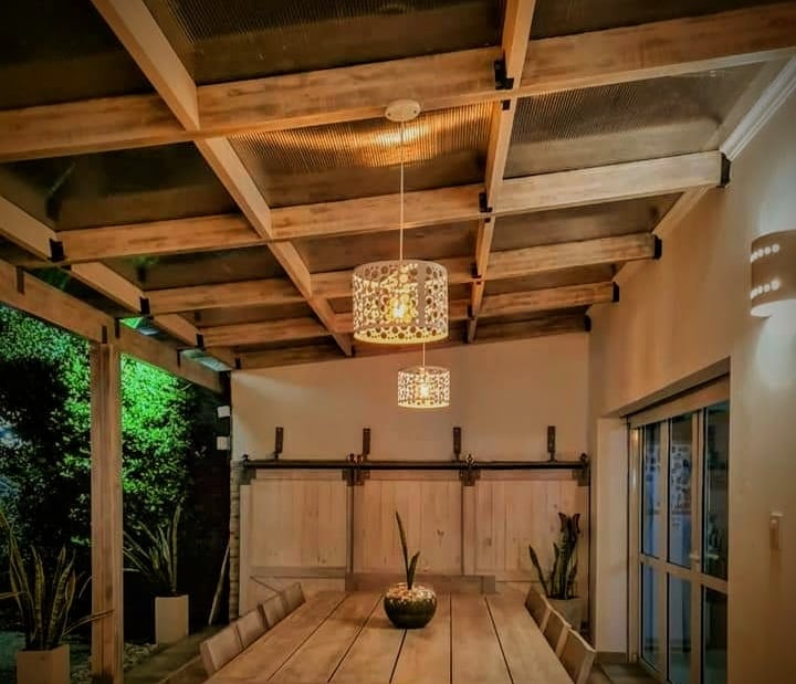 Comedor rústico al aire libre con mesa de madera, iluminación colgante y pérgola de madera.