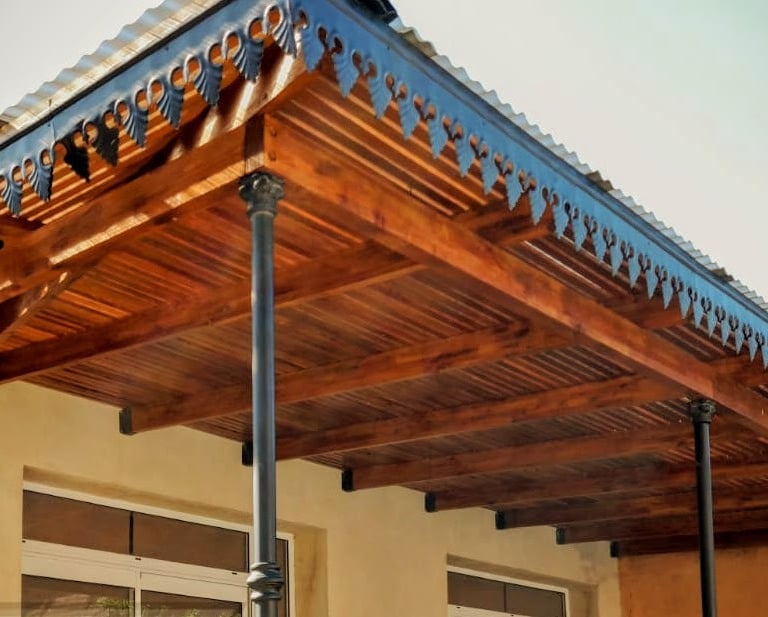 Toldo rústico de madera para porche con detalles decorativos de celosía y pilares de soporte negros.